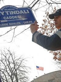 Pracovník městského úřadu u cedule s novým jménem ulice; v pozadí vlajka USA na střeše americké ambasády.